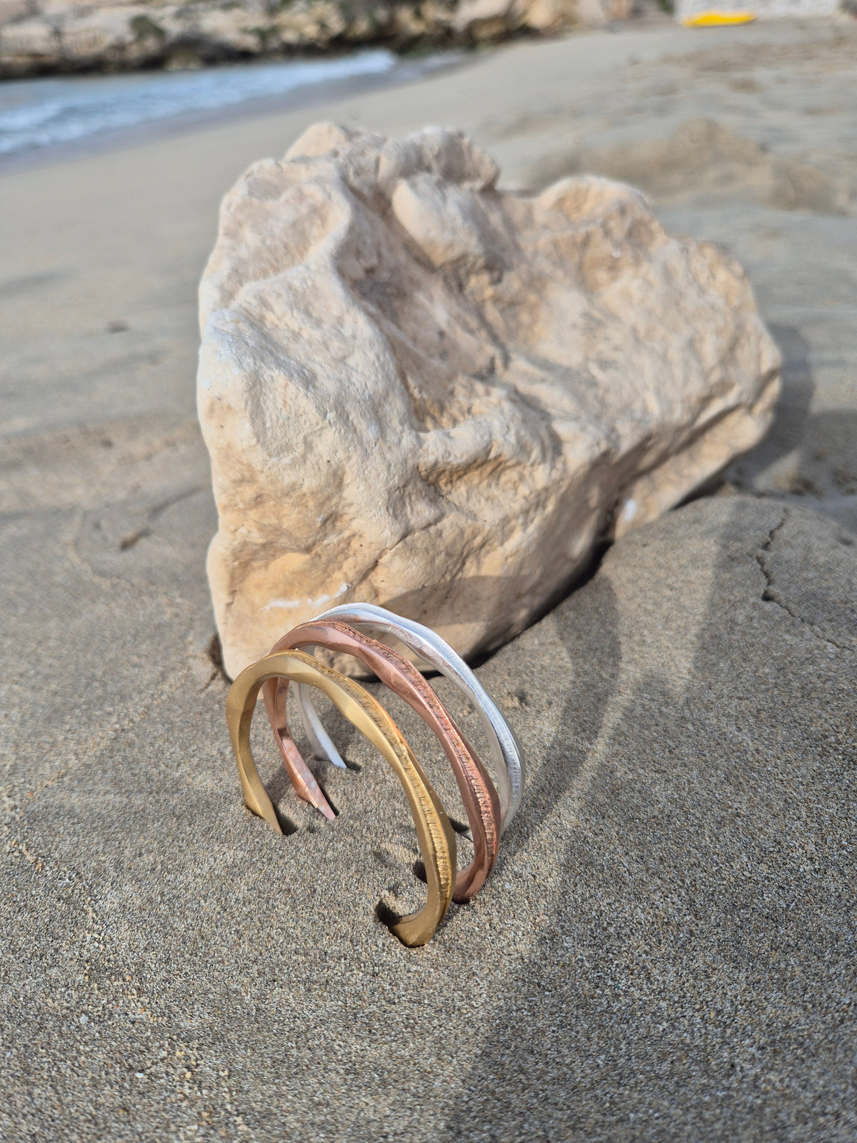 Tre bracciali Anima Tiziana Piola in finitura dorata, oro rosa e argento appoggiati sulla sabbia